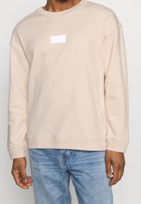 Light beige crew neck sweatshirt in soft fabric; features a small white rectangular logo on the chest; relaxed fit with long sleeves.