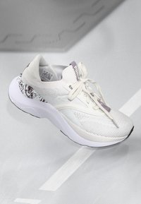 Chaussure de sport blanche avec un dessus en tricot texturé, des accents violets et une semelle rembourrée. Le talon présente un revêtement à motifs unique.