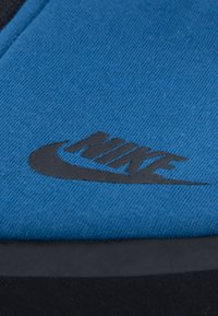 Zila auduma virsma ar melnu Nike logotipu. Gluda tekstūra ar kontrastējošu melnu zoli. Apaļa forma, paredzēta ikdienas valkāšanai.