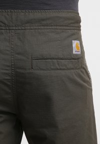 Mørkegrønne bukser med et ternet mønster, der har en baglomme og et hvidt lærreds-patch med en gul logo. Elastisk linning for komfort.