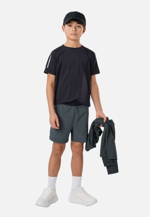 Chłopiec w czapce z daszkiem w kolorze czarnym, czarnej koszulce, szarych szortach, białych skarpetkach i sportowych butach, trzymający w prawej ręce złożoną ciemną kurtkę.