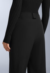 Černé kalhoty na míru s hladkým povrchem, širokým pasem a jemnými švy. Materiál je pružný a těsně přiléhající.