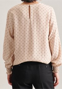 Blusa in tessuto beige con pois bordeaux, maniche a sbuffo e un piccolo dettaglio a chiave sulla schiena. Polsini testurizzati che si stringono ai polsi.