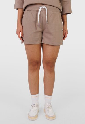 Persona che indossa pantaloncini beige con coulisse, calzini bianchi con scritta arancione e sneakers bianche, in piedi contro uno sfondo chiaro e uniforme.