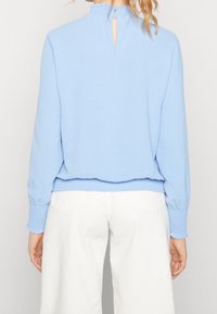 Maglione azzurro chiaro con finitura testurizzata, polsini arricciati e dettaglio a chiave sul retro. Abbinato a pantaloni bianchi con tasche posteriori.