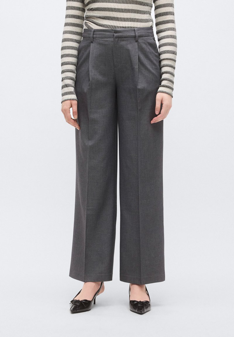 Παντελόνια με φαρδιά πόδια σε σκούρο γκρι ύφασμα με ραμμένη εφαρμογή, διαθέτοντας πτυχώσεις στο μπροστινό μέρος, λεία υφή και ψηλή μέση. Τονίζονται με κλασικά τακούνια.