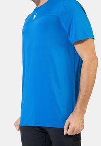 Camiseta deportiva azul con una textura suave, con cuello redondo, mangas cortas y un pequeño logo de araña en el pecho.