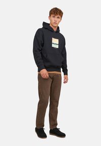 Schwarzer Kapuzenpullover mit einer Fronttasche und Grafikdruck. Kombiniert mit braunen Hosen und schwarzen Schuhen mit strukturiertem Profil.