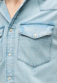 Ανοιχτό μπλε τζιν πουκάμισο με κουμπωτές τσέπες στο στήθος και μεταλλικά κουμπιά παρόμοια με τρουκς κατά μήκος της εμπρόσθιας λωρίδας.