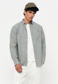 Camicia azzurra a maniche lunghe con strisce gialle verticali, colletto button-down e tasca frontale. Indossata sopra una maglietta bianca.