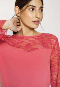 Top rosa com um corpo de tecido liso e mangas de renda transparente com padrões florais e um colarinho arredondado.