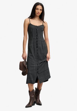 Mujer con vestido midi sin mangas, de estampado en blanco y negro, sosteniendo un sombrero marrón y llevando botas marrón oscuro hasta media pantorrilla.