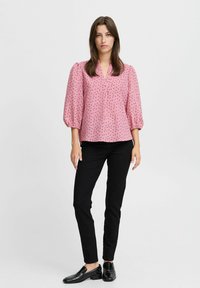 Rosa Bluse mit schwarz geometrischen Mustern, puffärmelig und mit V-Ausschnitt. Kombiniert mit schwarzen, figurbetonten Hosen und glänzenden schwarzen Slippers.
