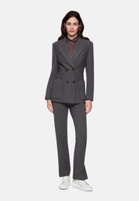 Gray double-breasted suit with textured fabric, side pockets, and matching trousers. Paired with a patterned shirt and white sneakers.