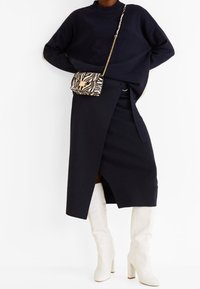 Granatowy sweter z dzianiny o wysokim kołnierzu, czarna spódnica o kroju kopertowym oraz białe kozaki za kolano na obcasie. Torebka w paski zebry dodaje kontrastu.