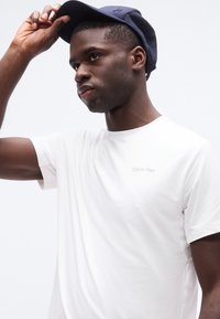 Νέος άνδρας που φοράει λευκό μπλουζάκι Calvin Klein ρυθμίζει το σκούρο μπλε καπέλο του μπροστά σε απλό λευκό φόντο.