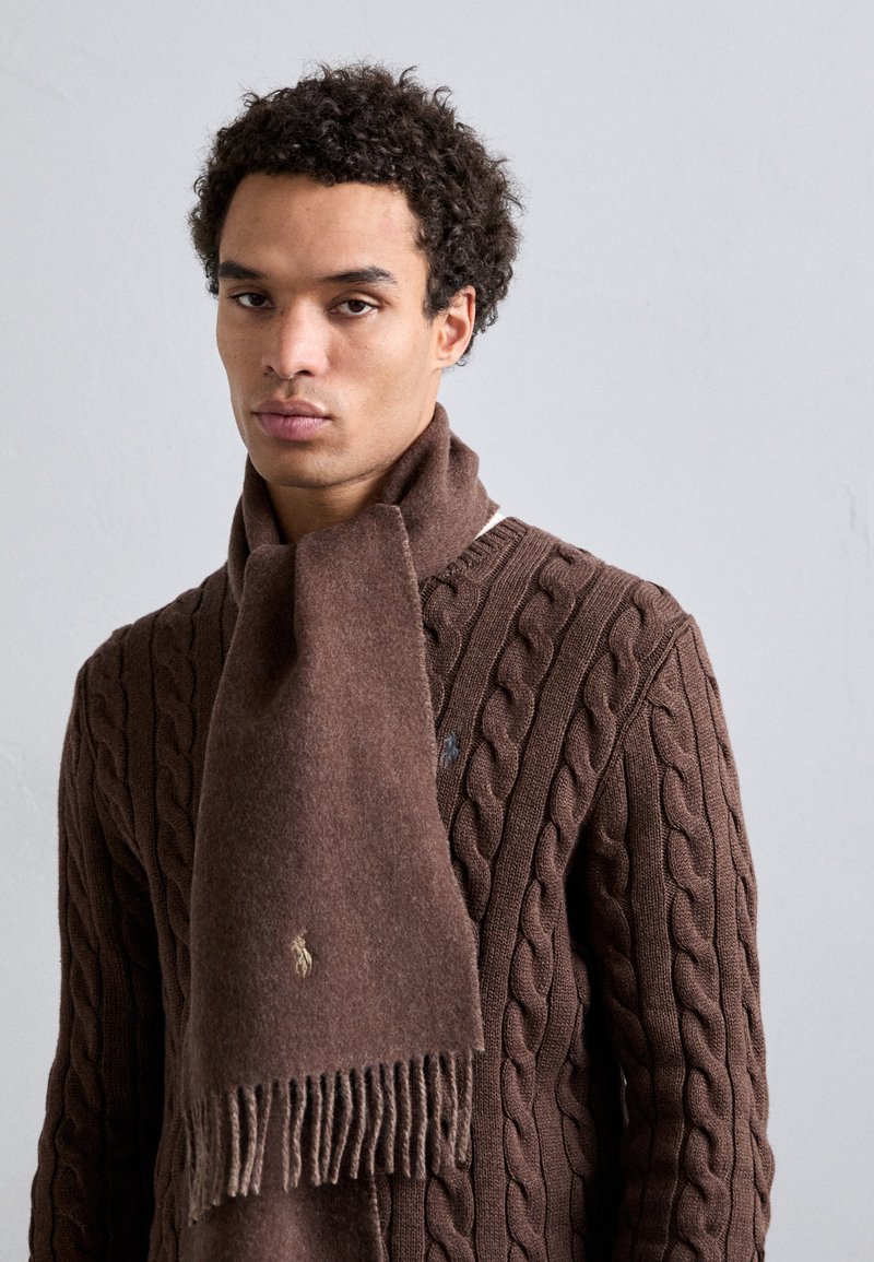 Καφέ μάλλινο κασκόλ με φραμπαλάδες, φορτωμένο πάνω από ένα υφαντό καφέ πουλόβερ με πλέξη καλωδίου. Διακριτική λεπτομέρεια με λογότυπο ορατή στο κασκόλ.