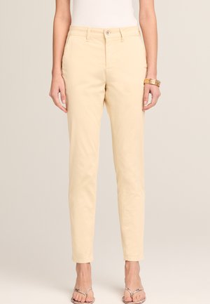 Frau trägt beige gerade geschnittene Hosen, weißes Oberteil, goldene Armbänder am rechten Handgelenk und beige Sandalen mit Absatz, steht vor einfarbigem Hintergrund.