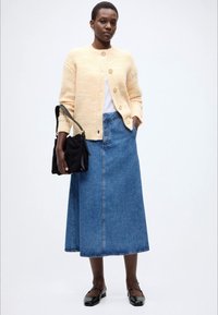 Κίτρινη ελαφρώς ανάγλυφη ζακέτα, λευκή μπλούζα, μακριά τζιν φούστα, μαύρη τσάντα χειρός και μαύρα flats. Λεπτή υφή και άνετη σιλουέτα.