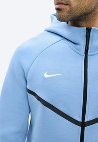 Άνδρας που φοράει ανοιχτό μπλε φούτερ Nike με φερμουάρ και μαύρη διαγώνια ρίγα, με ορατό κάτω μέρος του προσώπου και κοντή γενειάδα, σε λευκό φόντο.