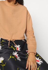 Lichtbruine cropped sweatshirt met een hoge hals, voorzien van een trekkoord in de taille, gecombineerd met een donkere bloemenrok met witte en rode madeliefjes.