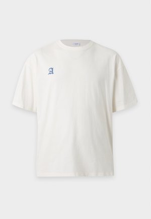Άτομο με πλεξούδες που φοράει λευκό t-shirt με μπλε "Pukka Copenhagen" κείμενο στην πλάτη και παντελόνι ελιάς, πάνω σε μονόχρωμο φόντο.