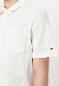 Homme portant un polo blanc texturé à manches courtes avec une poche avant et un petit logo sur la manche gauche.