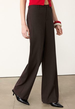 Mannequin portant un pantalon large marron foncé, des talons noirs à bout pointu, un top court rouge et des bracelets rouges et blancs au poignet gauche.