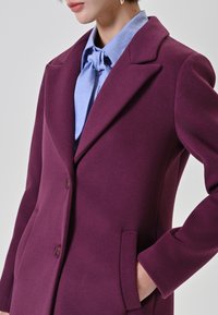 Donna che indossa un cappotto su misura viola con bottoni e tasche laterali, sopra una blusa azzurro chiaro annodata a fiocco al collo.