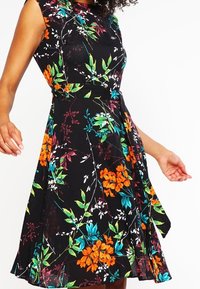 Robe sans manches noire avec un motif floral vif en orange, vert, bleu et blanc, dotée d'une taille cintrée et d'une jupe évasée.
