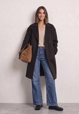 Femme aux cheveux mi-longs portant un manteau sombre, un pull beige, un jean à jambes larges et des chaussures noires, tenant un sac à main en daim marron.