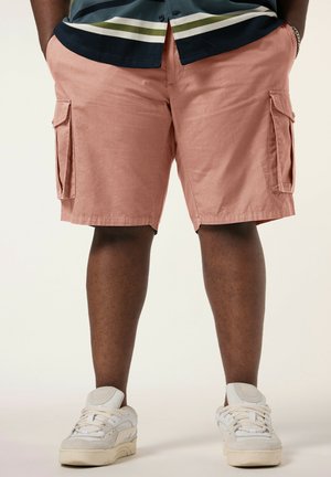 Mann trägt lachsrosa Cargo-Shorts, weiße Sneakers und ein dunkles Hemd mit grünen und weißen Streifen, steht mit den Händen in den Taschen.