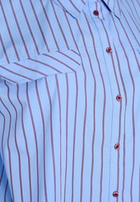 Lysblå skjorte med vertikale røde striber. Har spidse krave, brystlomme og kontrastknapper.