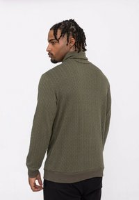 Pull-over en tricot vert olive avec un col haut, featuring un motif texturé. Des poignets et un ourlet côtelés ajoutent de la structure. Modèle vu de dos.