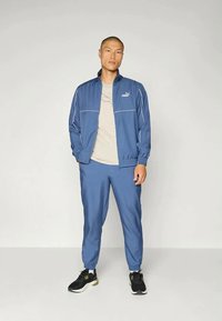 Blauwe trainingsset van soepel, lichtgewicht materiaal. Zip-up jacket met witte accenten, bijpassende tapered broek, en zwarte sportschoenen.