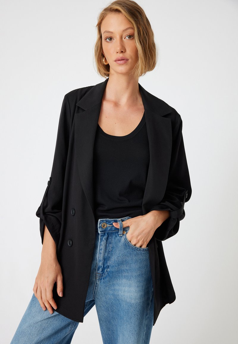 Chaqueta negra de corte relajado, con grandes botones y mangas enrolladas, combinada con una camiseta de tirantes negra y jeans de tiro alto azul claro.