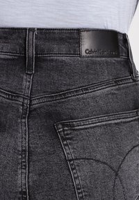 Pantalones vaqueros de mezclilla negros con una textura suave, que presentan un parche de cuero en la cintura con la etiqueta "Calvin Klein Jeans" y diseño estándar de bolsillos.