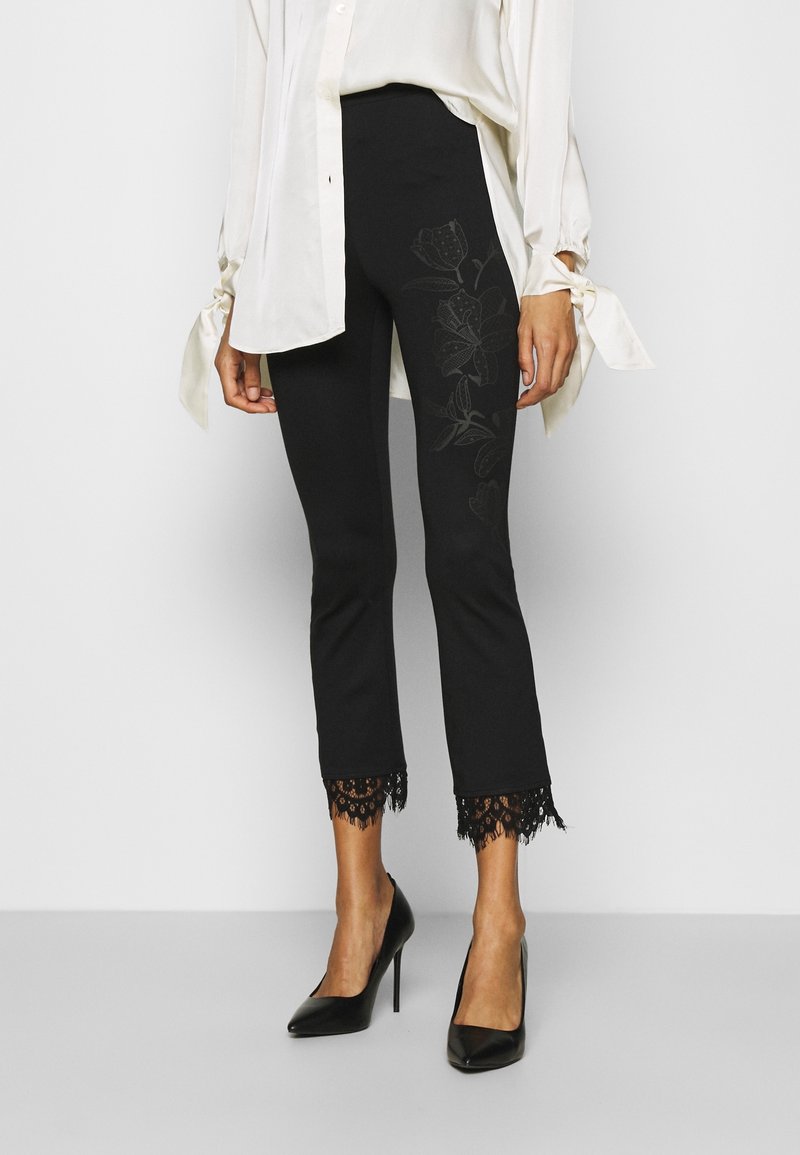 Zwarte leggings met een bloemenpatroon aan de zijkant, cropped lengte en kantafwerking aan de zoom. Gecombineerd met een witte blouse en zwarte hakken.