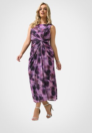 Kobieta ubrana w fioletową i czarną sukienkę midi bez rękawów z wzorem i skręcanym detalem na talii, w zestawieniu ze srebrnymi sandałami na wysokim obcasie.