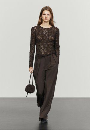 Donna che indossa un top nero in pizzo a maniche lunghe, pantaloni larghi marrone scuro, scarpe nere e porta una piccola borsa nera mentre cammina in avanti.