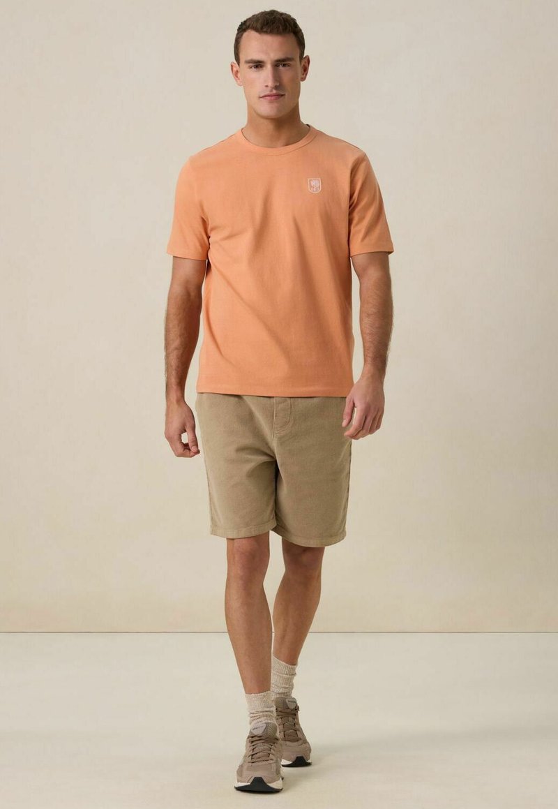 Korte mouw perzikkleurig katoenen T-shirt met een logo, gecombineerd met beige corduroy shorts, beige sokken en bruine sport schoenen.