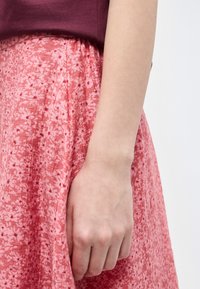 Αριστερό χέρι και χέρι ενός ατόμου που ακουμπούν δίπλα σε μια ροζ φλοράλ πλισέ φούστα και μαρόν μπλούζα σε απλό λευκό φόντο.