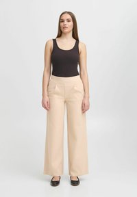 Top in maglia nera a costine abbinato a pantaloni beige a vita alta e gamba larga, con pieghe frontali e scarpe basse. Sfondo uniforme.