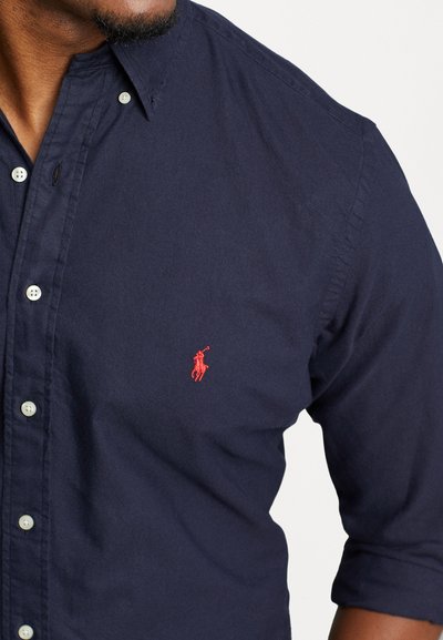 Chemise boutonnée bleu marine en coton, avec un logo rouge brodé sur la poitrine gauche ; comprend un col boutonné et des boutons blancs.