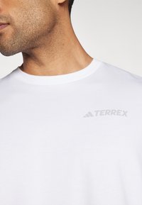 Camiseta blanca de algodón con cuello redondo y un pequeño logo gris "TERREX" en la parte superior izquierda. Textura suave y ajuste clásico.