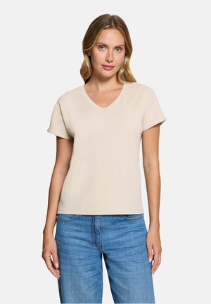 Femme aux cheveux blonds portant un haut beige à manches courtes avec un col en V et un jean bleu, debout devant un fond clair uni.