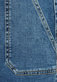 Denimkangas keskisinisena; siinä on silmiinpistävä tasku, jossa on kontrastoiva keltainen ommel ja pehmeä, teksturoitu pinta.