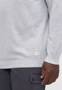Grauer Strickpullover mit strukturiertem Muster, gerippten Bündchen und Saum sowie einem kleinen Markenlabel am Saum. Kombiniert mit dunklen Hosen.