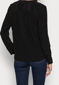Blusa nera a maniche lunghe con texture liscia, caratterizzata da un collo alto, cuciture sulle spalle discrete e polsini con bottoni.
