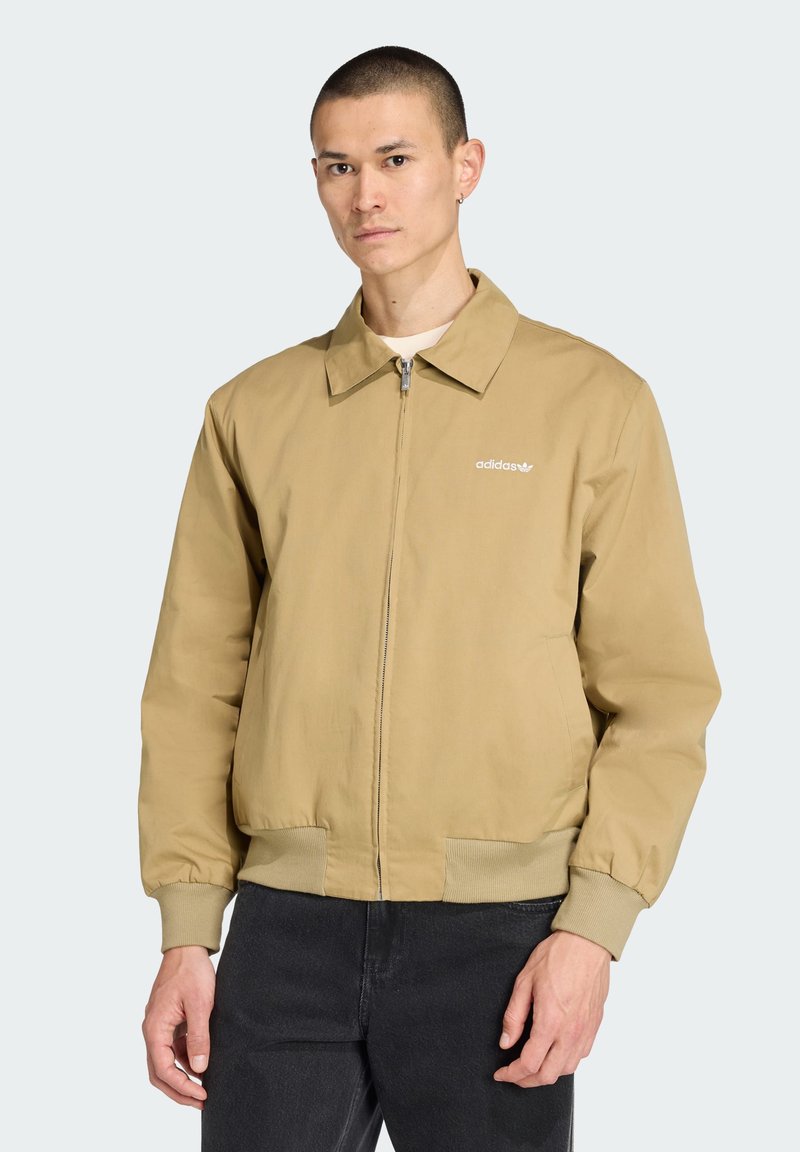 Jachetă bomber bej cu guler, fermoar frontal și manșete elastice. Prezintă logo-ul alb Adidas pe pieptul stâng.